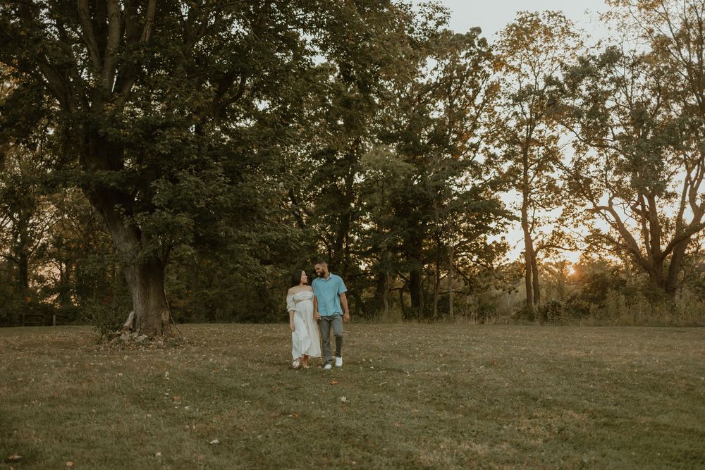 Couple walking together at sunset in a grassy field with tall trees silhouetted against the warm evening sky.