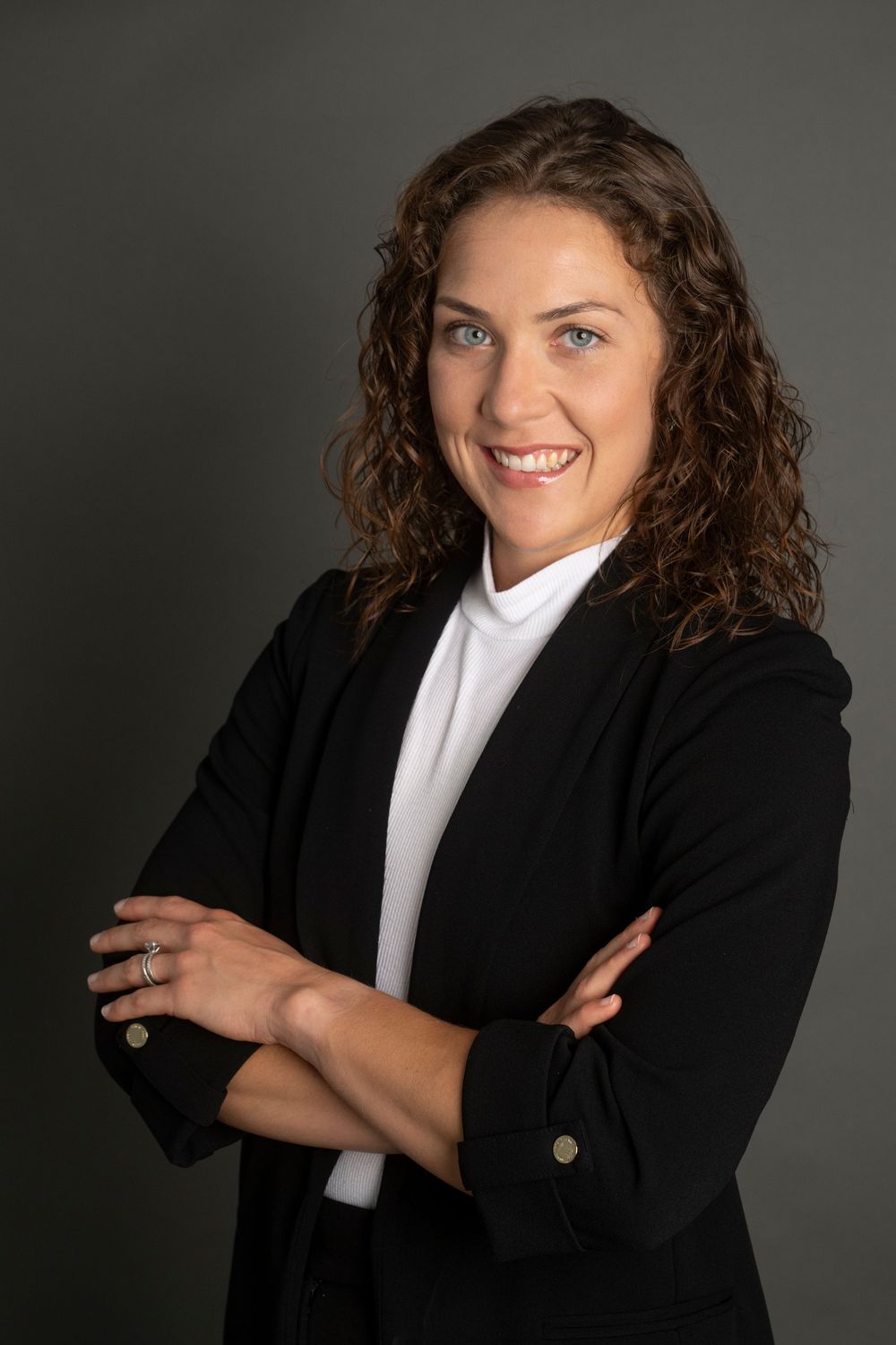Professional headshot of a person in a black blazer and white blouse posing with crossed arms against dark background.