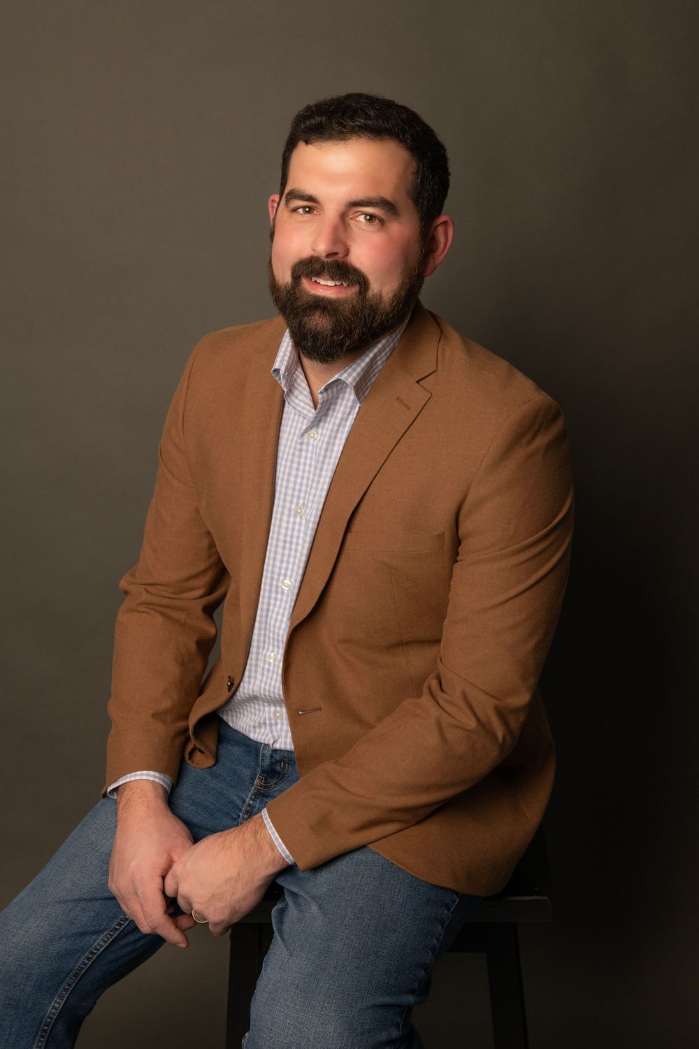 Professional studio portrait with brown blazer, gingham shirt and jeans against dark background.