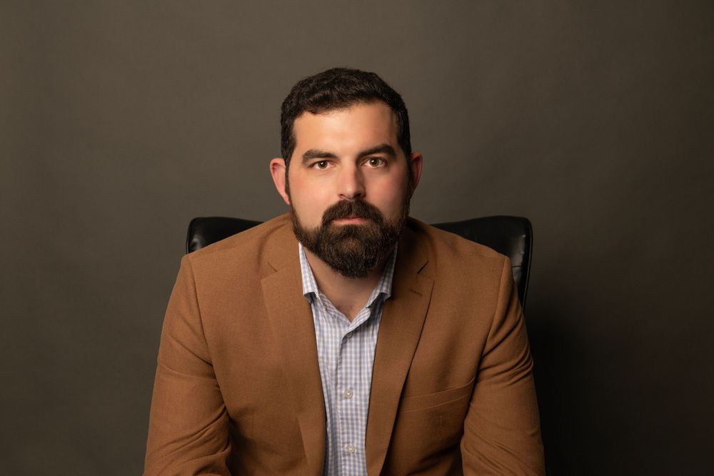 Professional headshot of person wearing a brown blazer and striped shirt against dark background.
