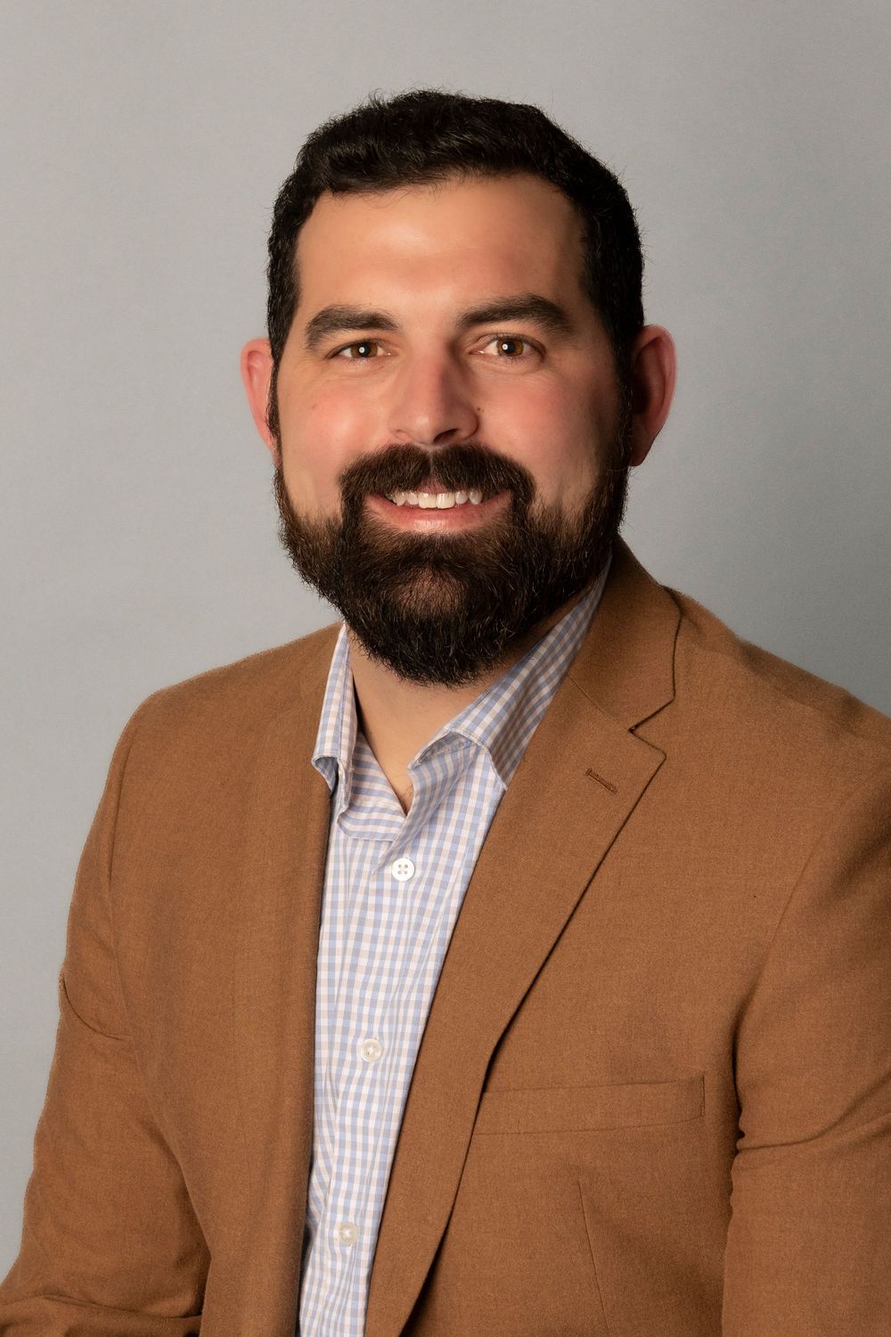 Professional business portrait of person wearing checkered button-down shirt and brown blazer against gray backdrop.