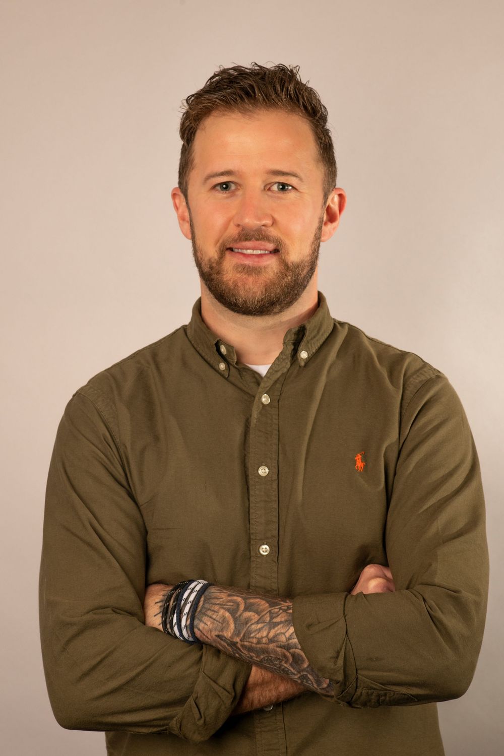 A headshot photo of someone wearing a brown button-down shirt with rolled sleeves and visible tattoos on their arm.