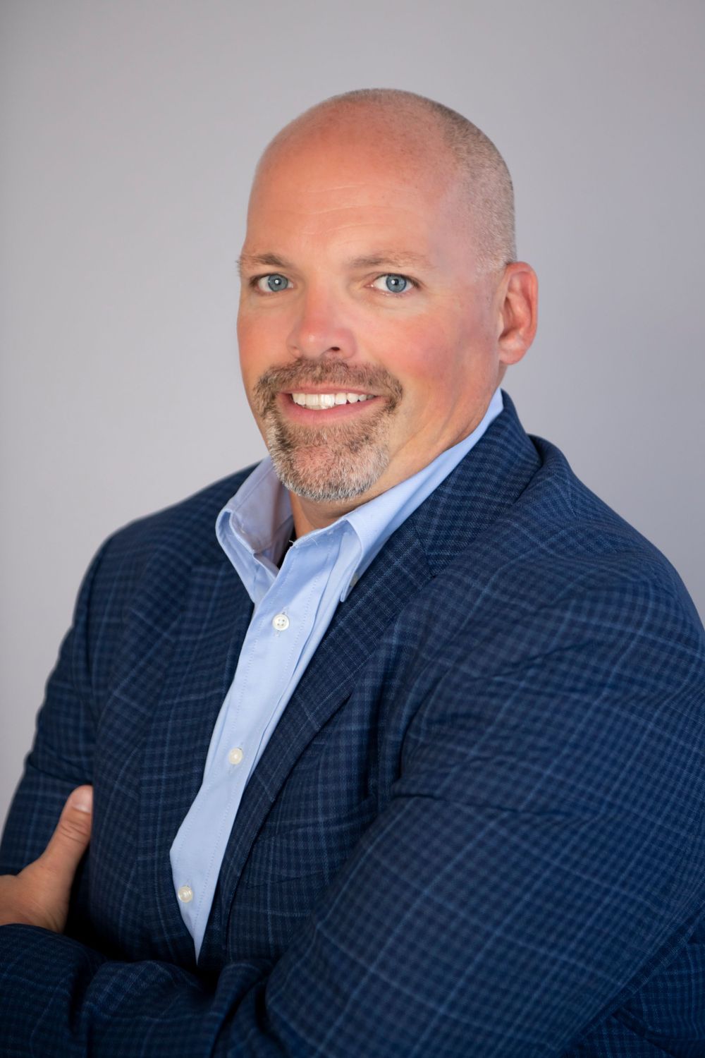 Professional headshot of person in navy checkered blazer and light blue shirt against grey background.