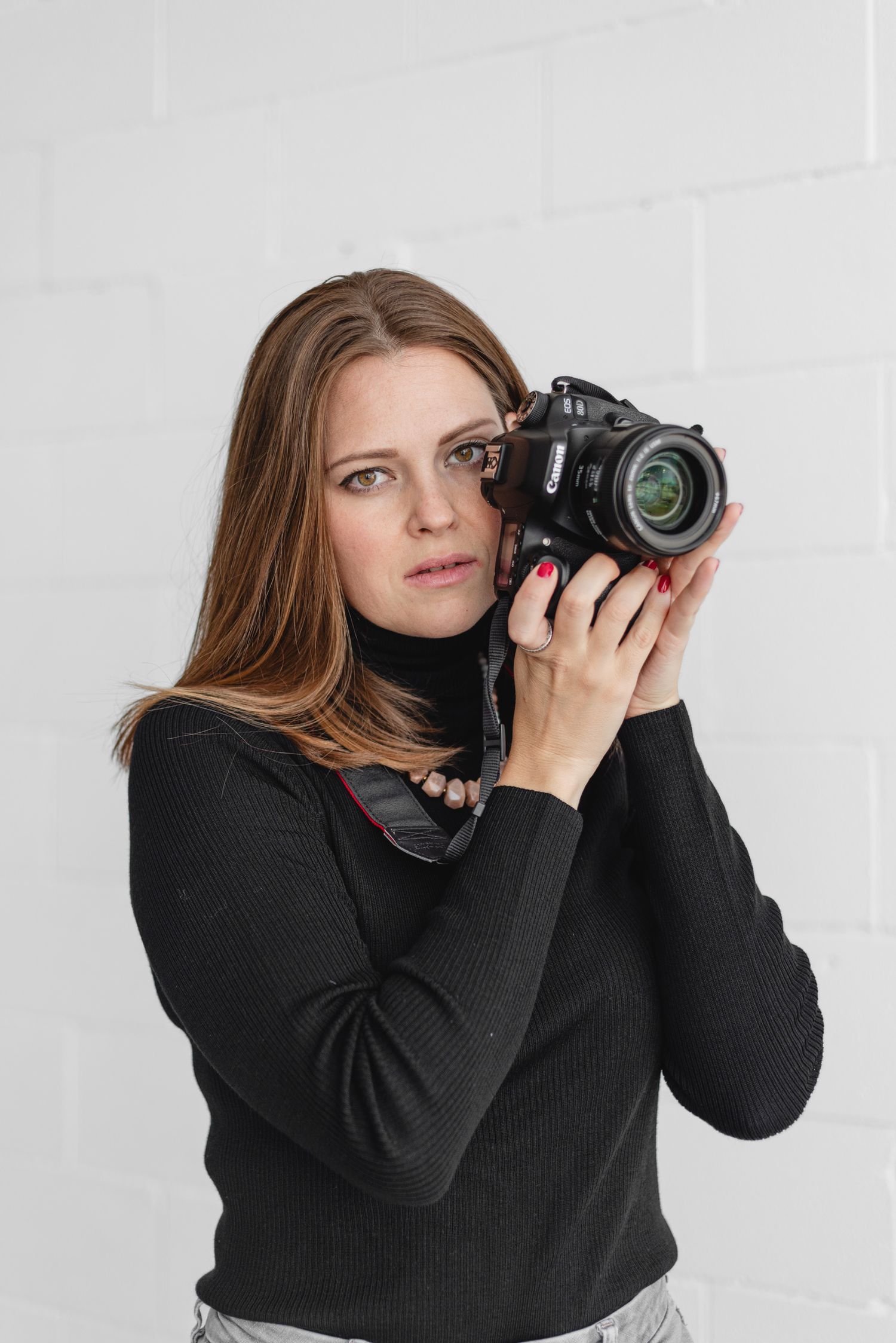 Photographer in black turtleneck holding DSLR camera against white wall.