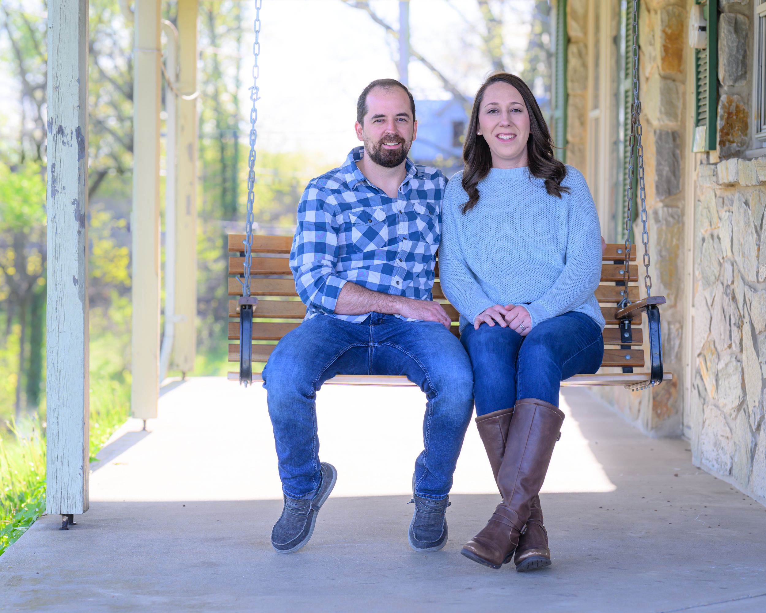 Couple sits together on wooden porch swing in front of stone wall with nature backdrop in casual attire.