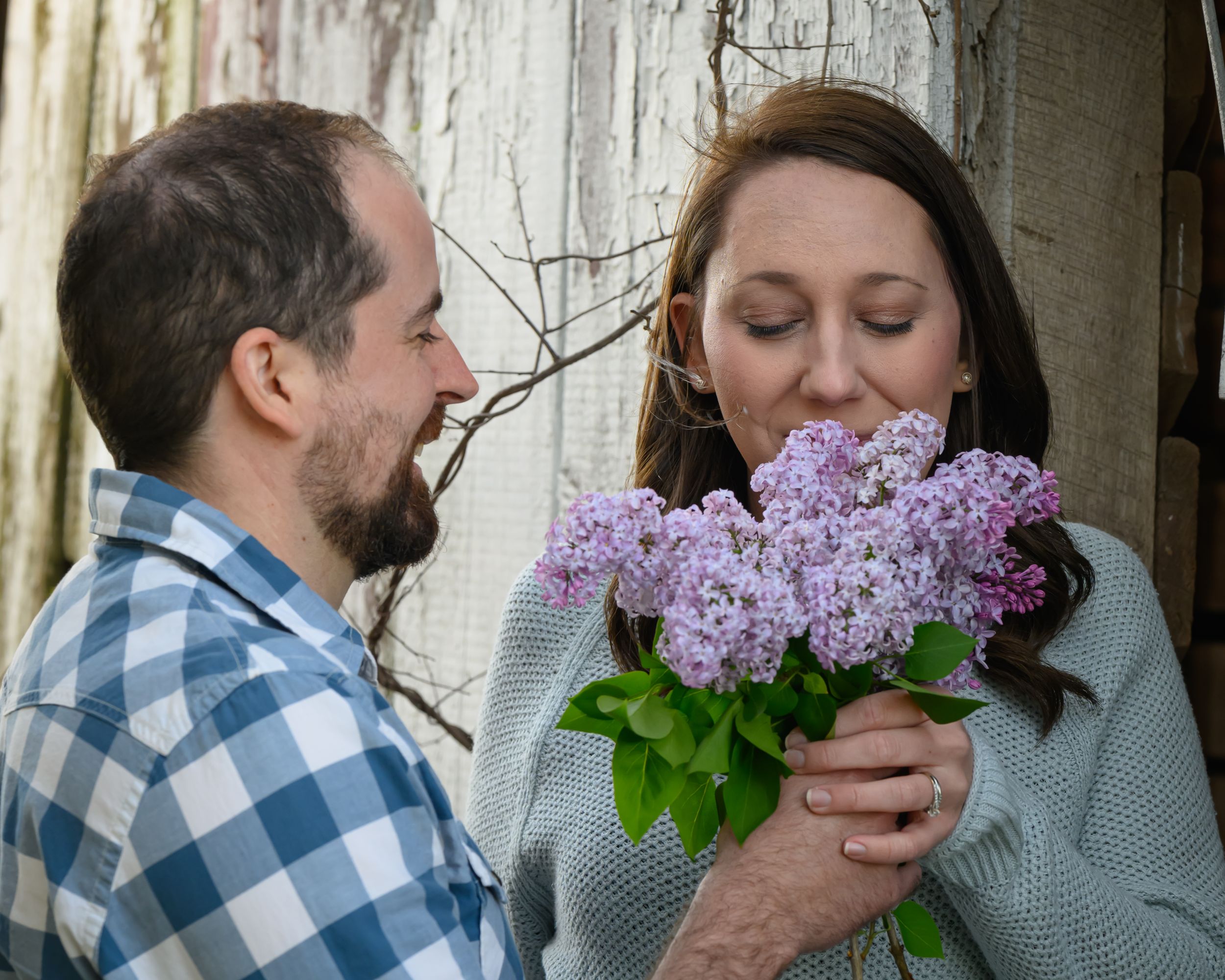 A couple share a tender moment outdoors as one smells a beautiful bouquet of purple lilac flowers.
