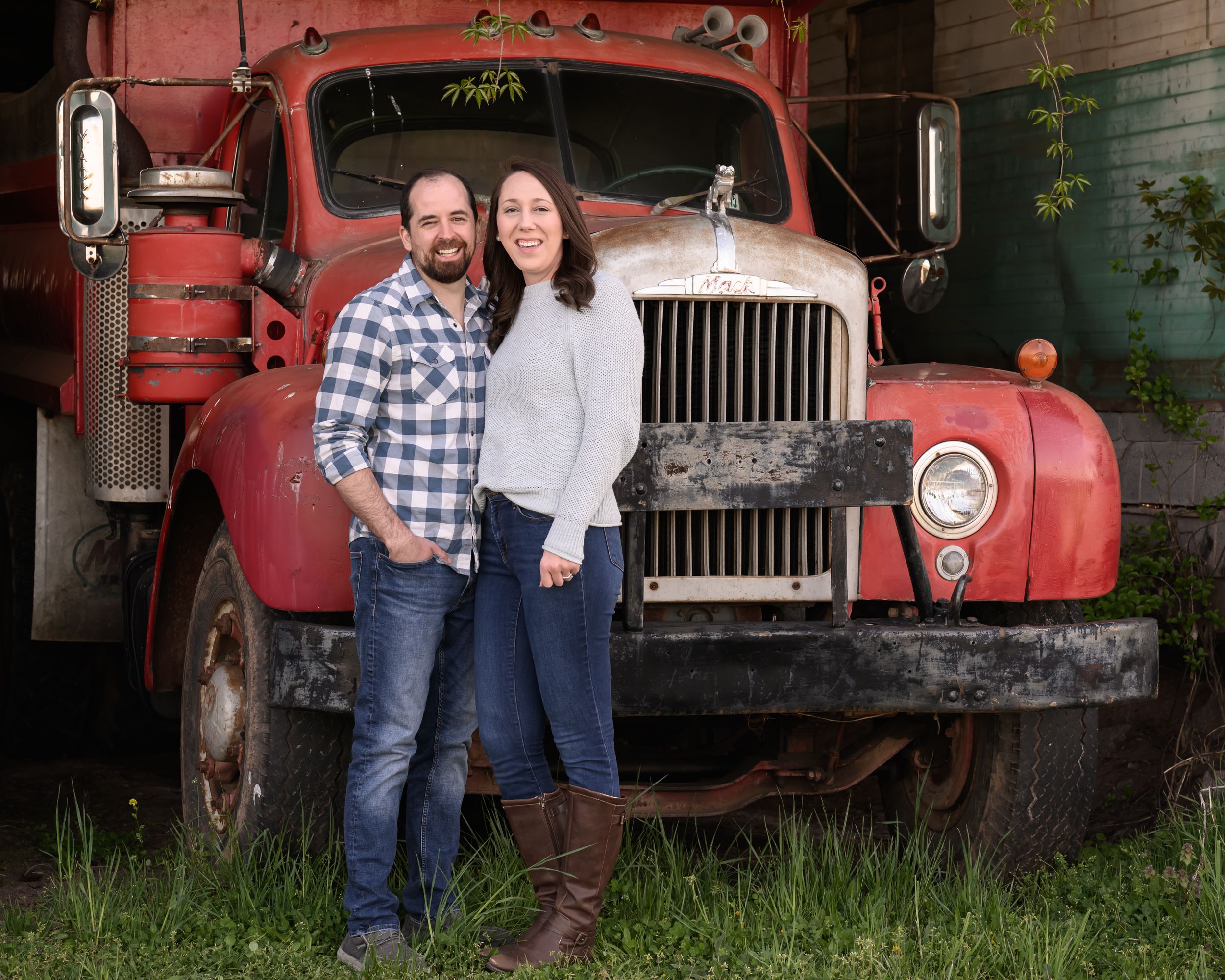 A couple poses together in front of a vintage red Mack truck on grass.