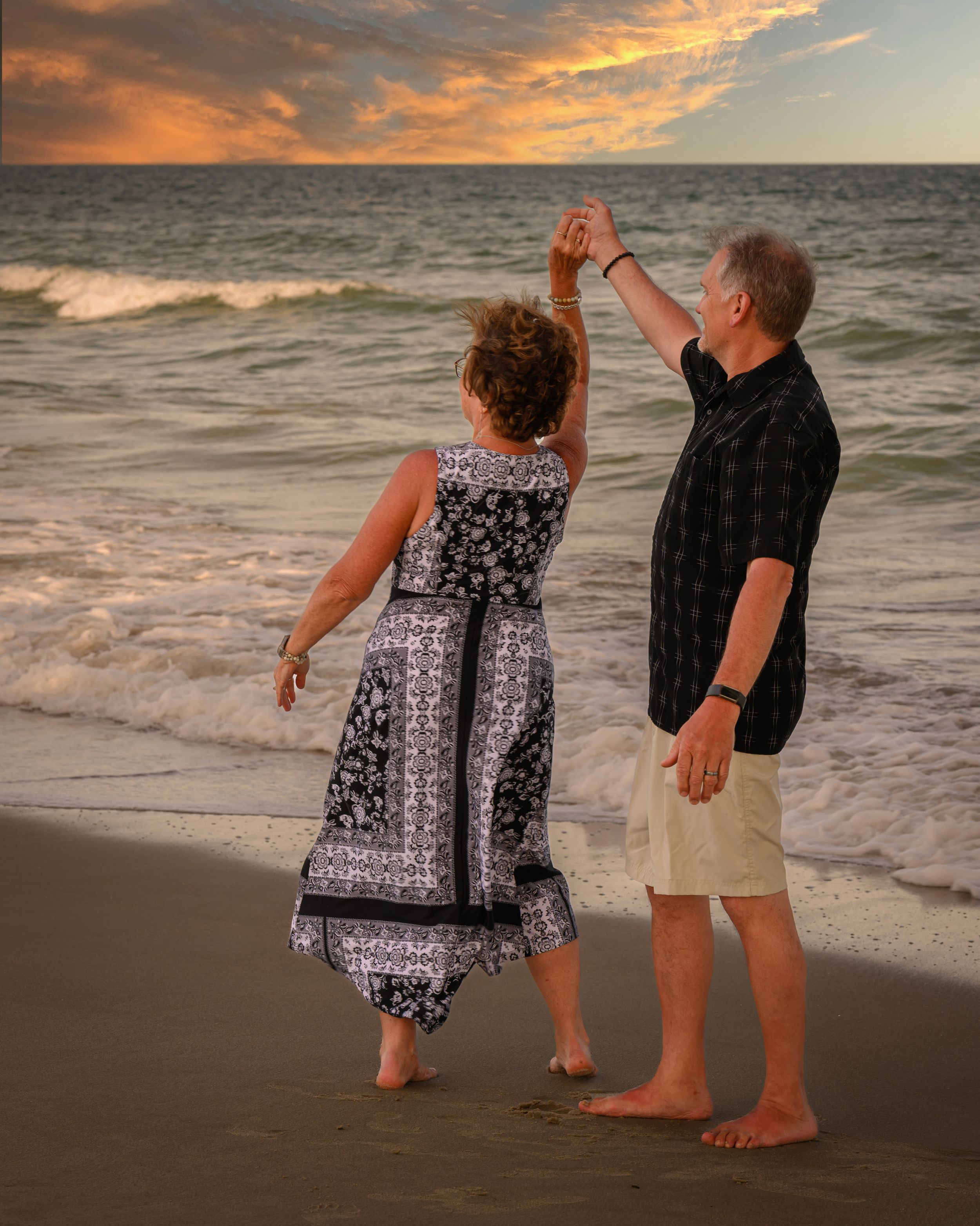 A couple dances barefoot on the beach at sunset while waves crash onto the shore and orange clouds fill the sky.