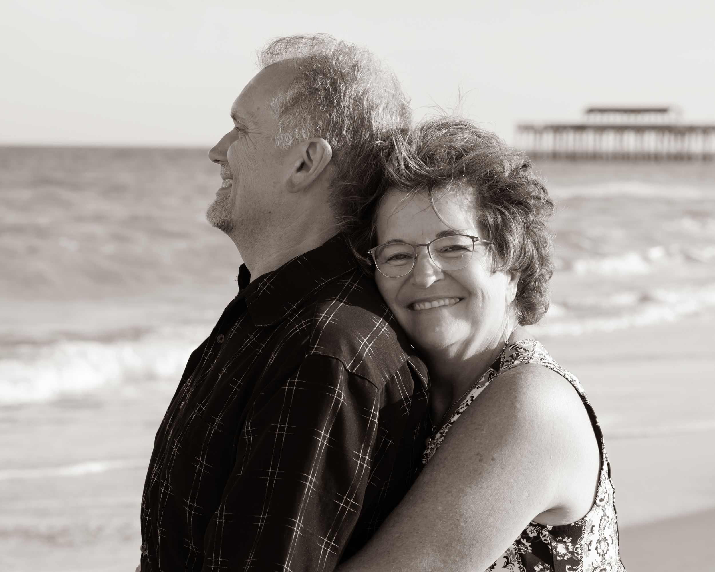 A tender moment captured in black and white of a couple embracing at the beach with a pier visible in the background.