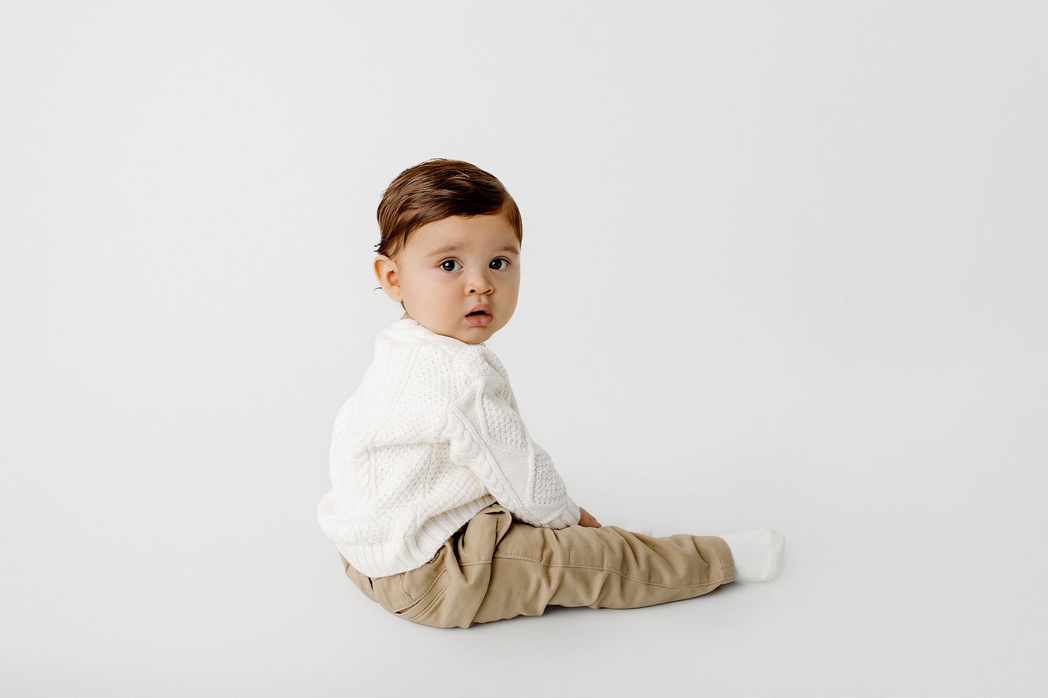 Adorable baby boy in white knit top and tan pants sitting on white background.