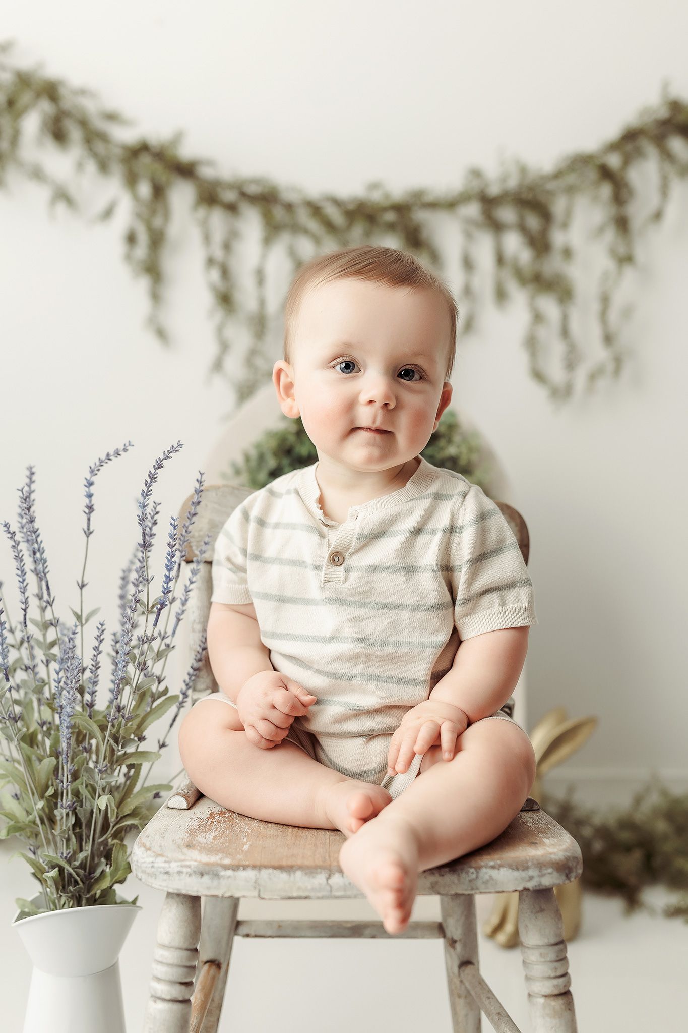 Sweet baby in striped onesie sits on vintage stool next to lavender bouquet under rustic greenery garland.