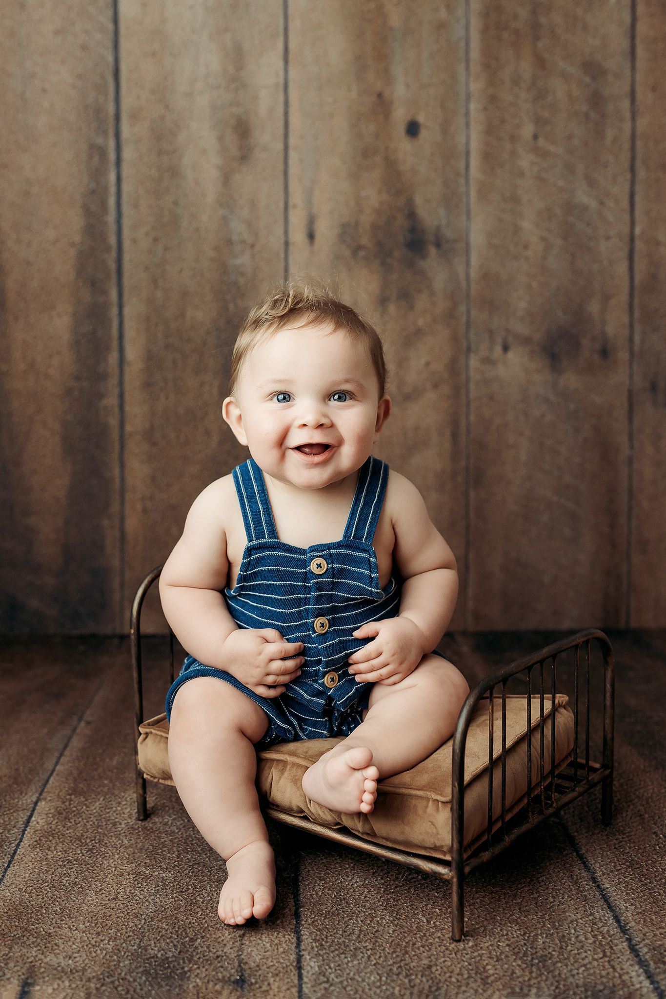 baby boy in striped overalls sits happily on a rustic metal bed frame against wooden wall backdrop.