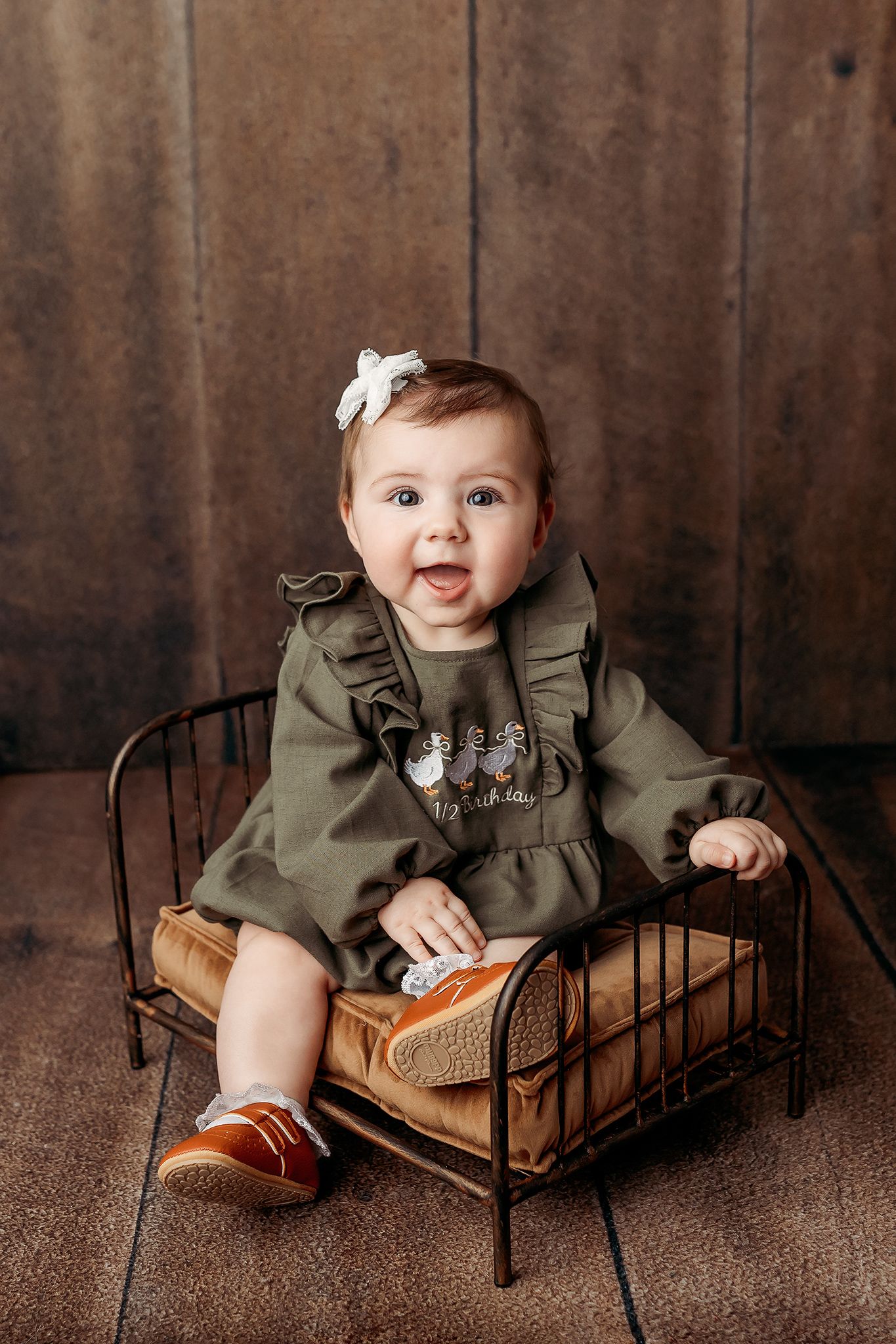 Smiling baby girl in green ruffle dress and white bow sits on a miniature vintage metal bed against a wood backdrop.