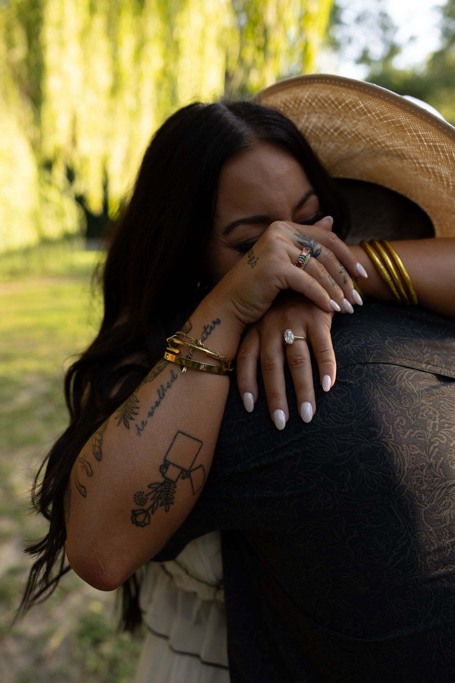 Woman with tattoos and gold bangles embracing a man wearing a cowboy hat under a willow tree.