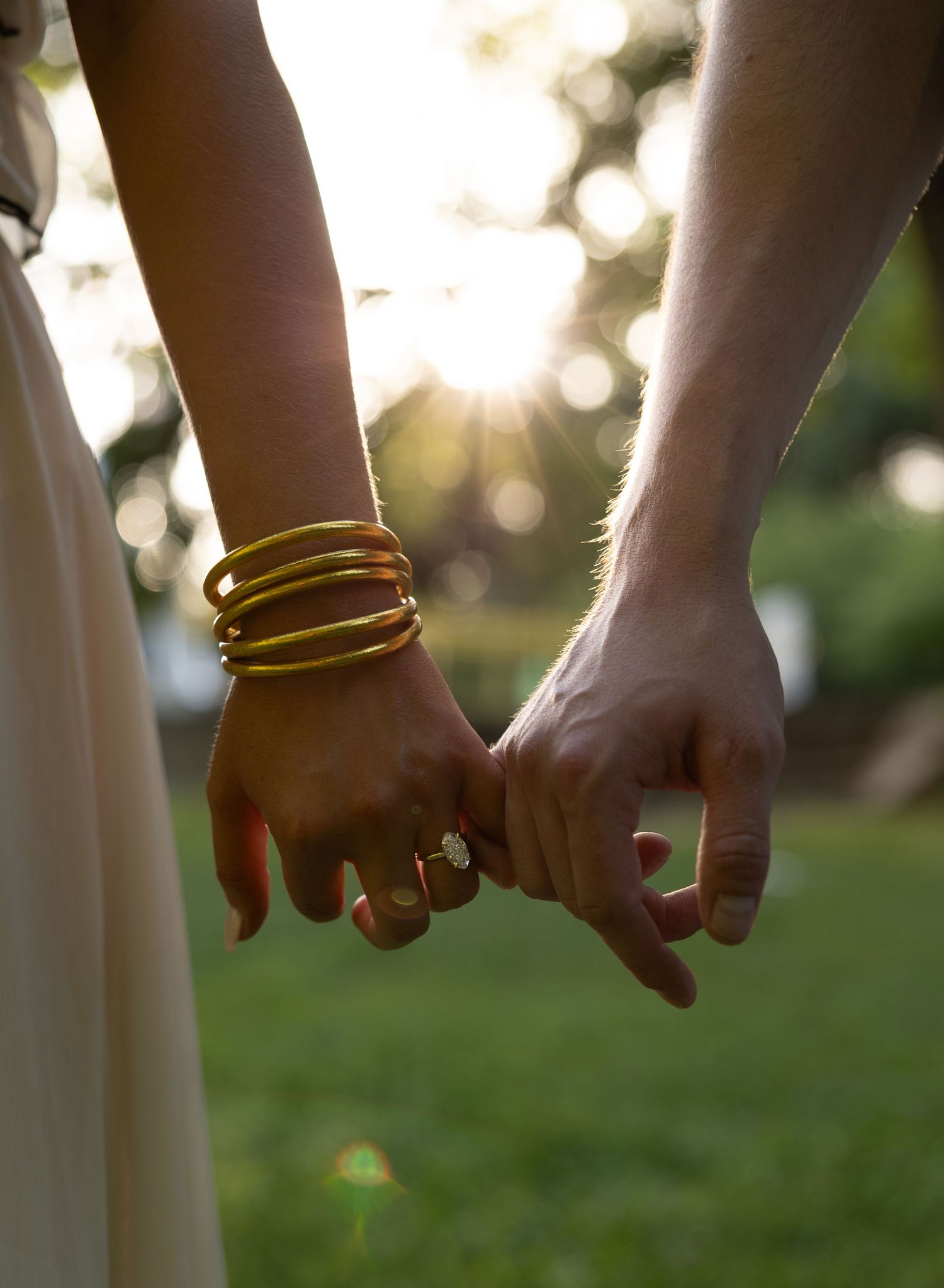 Couple holding hands with sunlight bokeh, woman wearing gold bangles and diamond engagement ring.