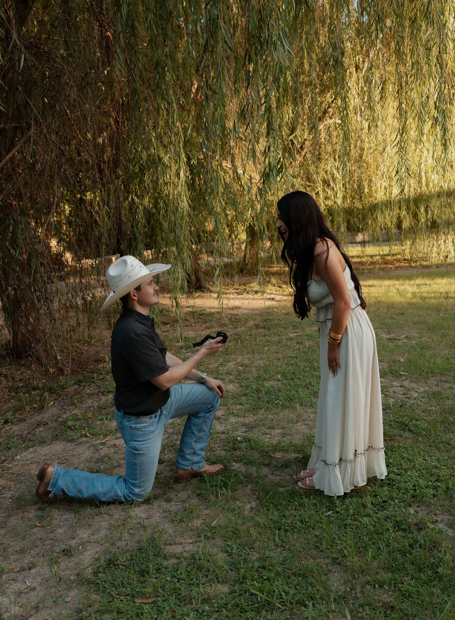 Man in cowboy hat kneeling to propose to woman in white dress under weeping willow tree.