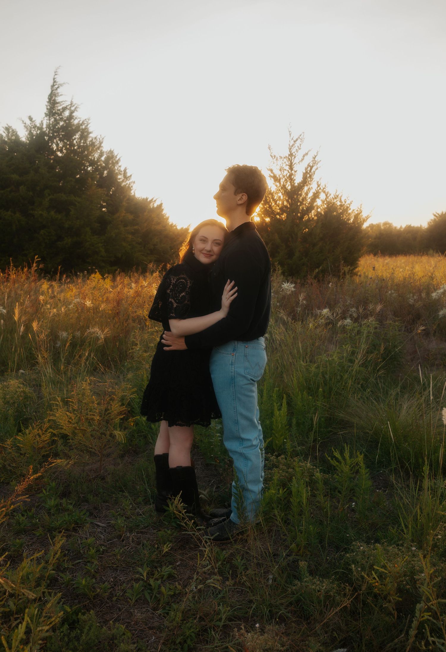 Couple embracing at golden hour sunset in a lush green field surrounded by trees.