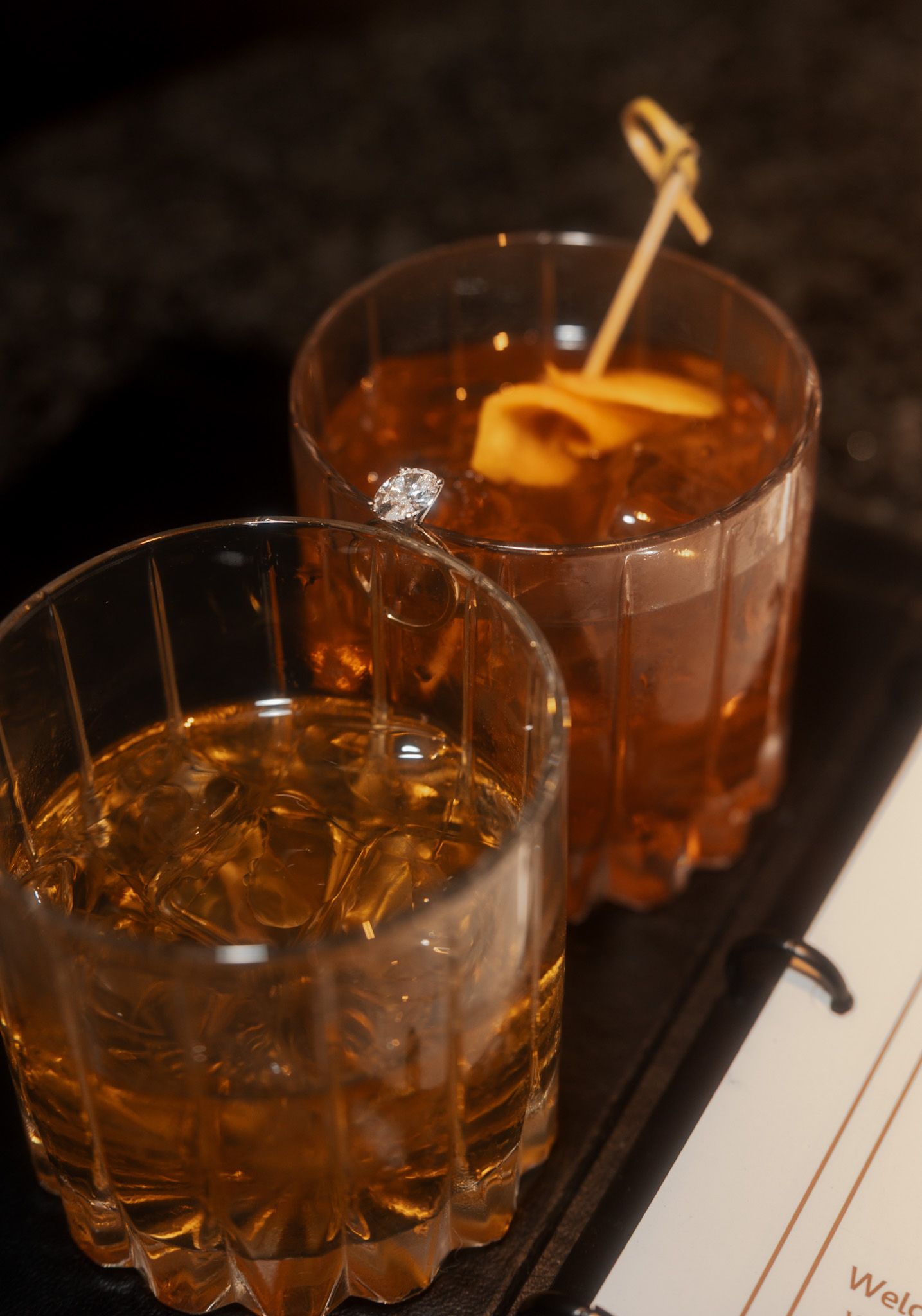Two whiskey cocktails on a dark bar tray, one featuring a diamond engagement ring as a surprise.