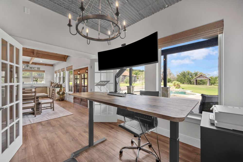 Modern home office with standing desk, mounted TV, circular chandelier, and French doors overlooking scenic outdoor view.