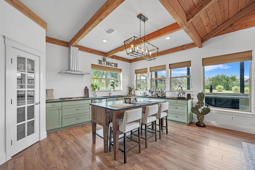 Modern farmhouse kitchen with sage green cabinets, wood beam ceiling, pendant lighting and large windows overlooking mountain views.