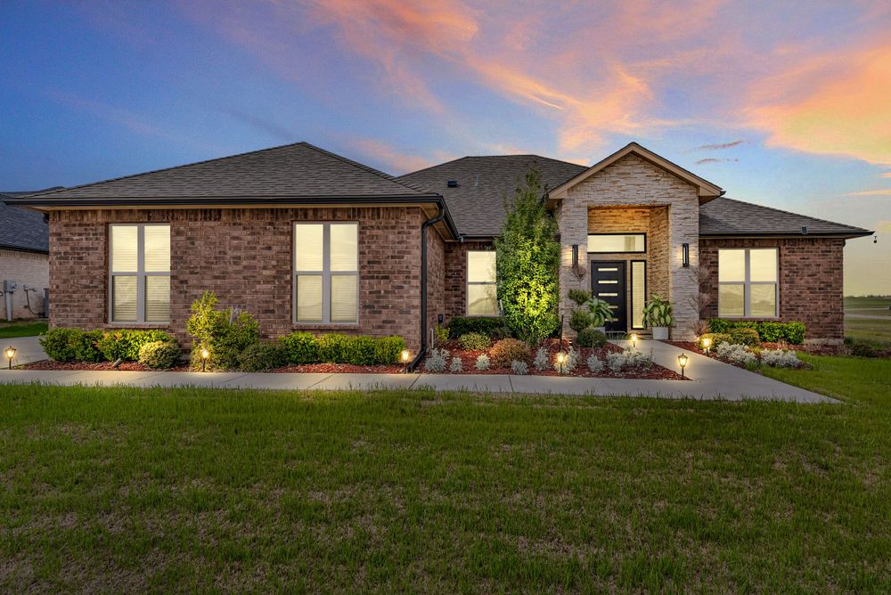 Modern ranch-style home with brick facade and decorative landscaping illuminated at sunset with warm glowing windows.