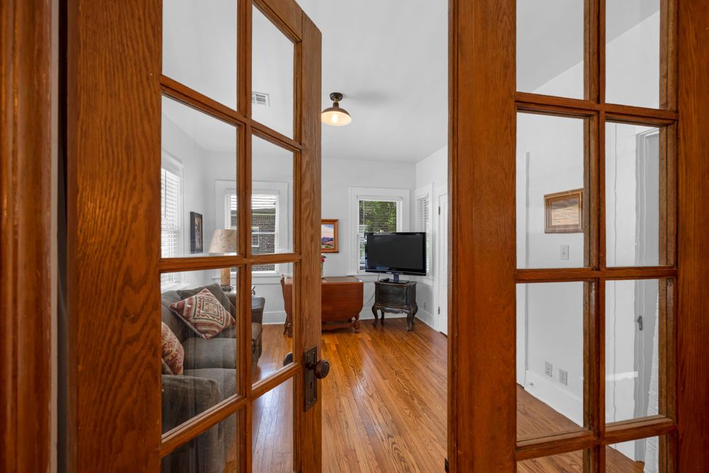 View through French doors into bright hallway with hardwood floors and natural woodwork leading to living space.
