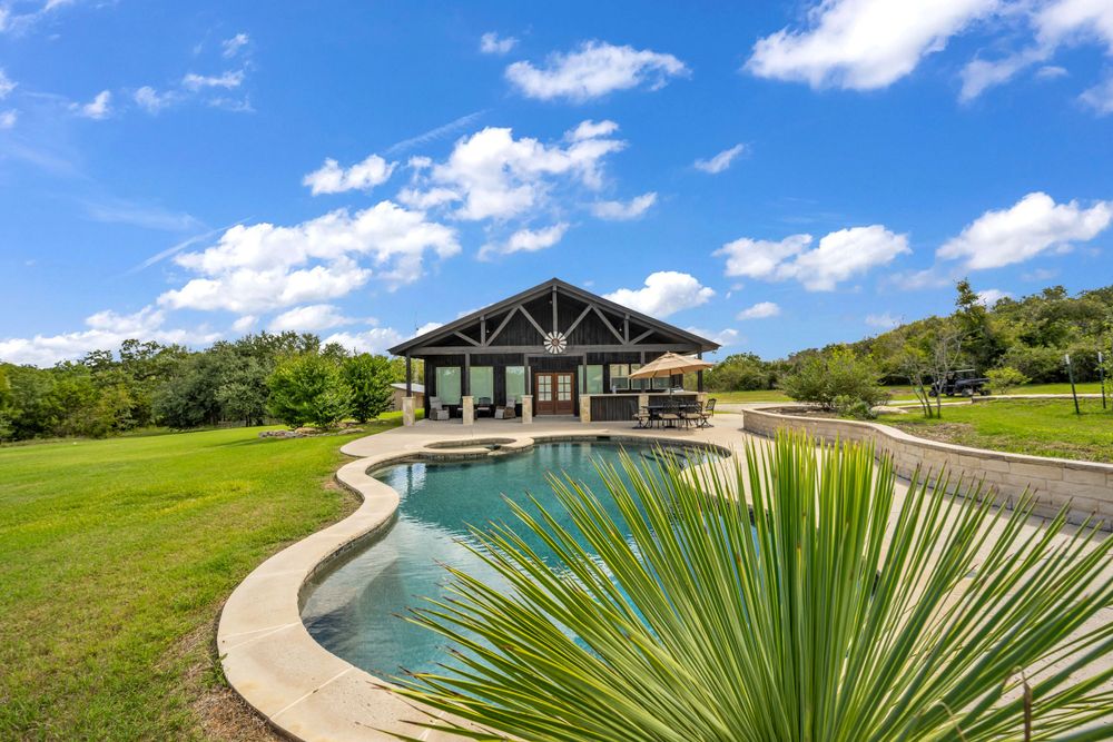 Luxury ranch house with curved swimming pool and palm plant surrounded by green hills under blue sky with white clouds.