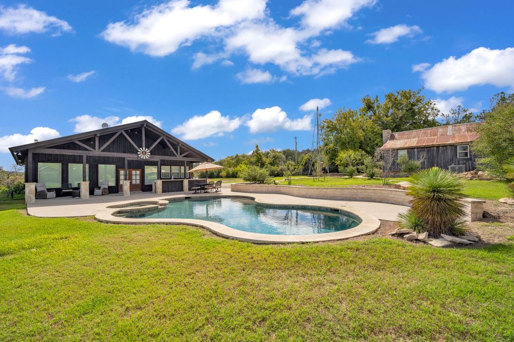 Luxurious backyard swimming pool and covered patio area surrounded by green lawn on a sunny day with blue skies.