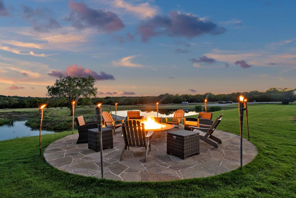 A cozy outdoor fire pit patio with Adirondack chairs and tiki torches overlooks a scenic lawn and pond at sunset.