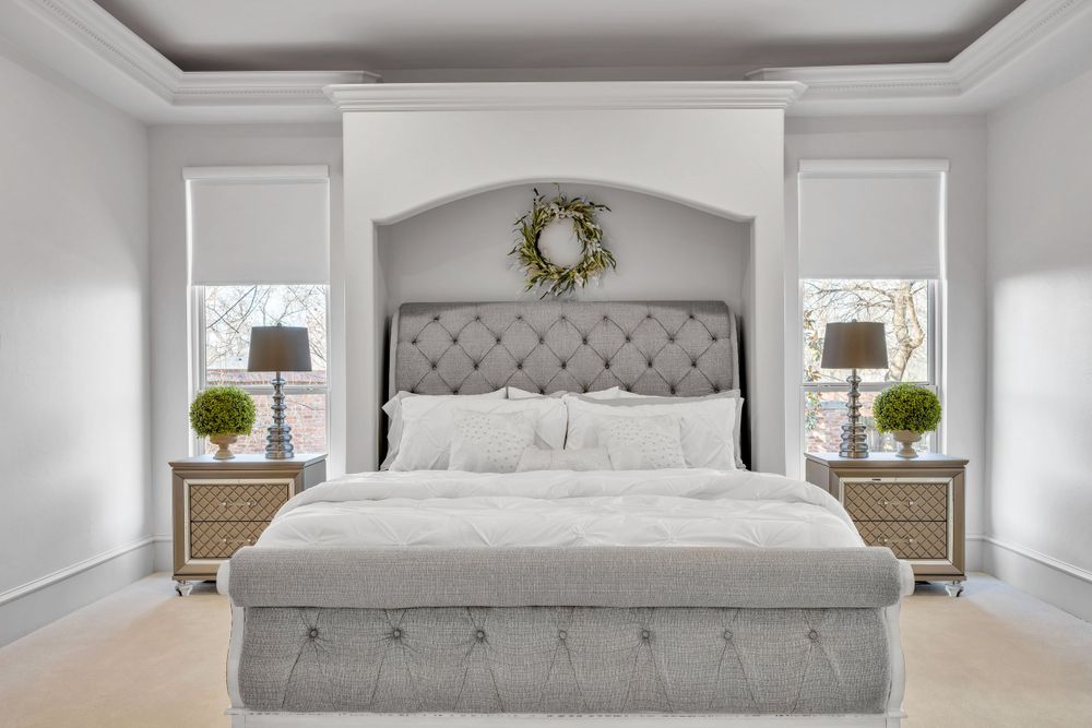 Elegant master bedroom with gray tufted headboard, matching nightstands, white bedding, and decorative wreath above.