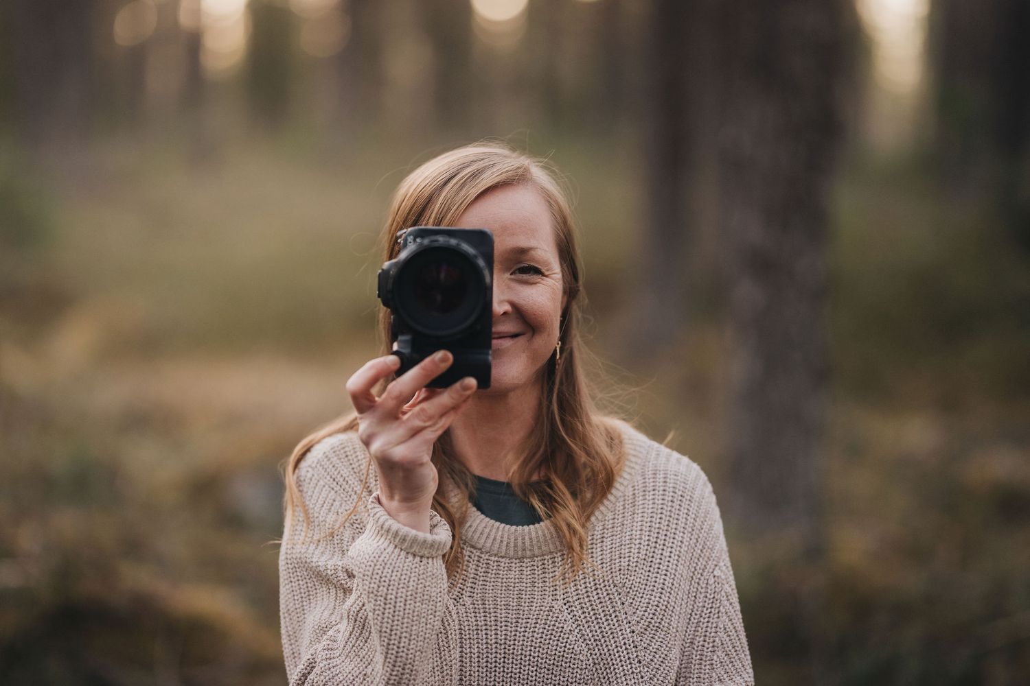 A smiling blonde woman holds a camera up to her face while standing in a softly lit forest.