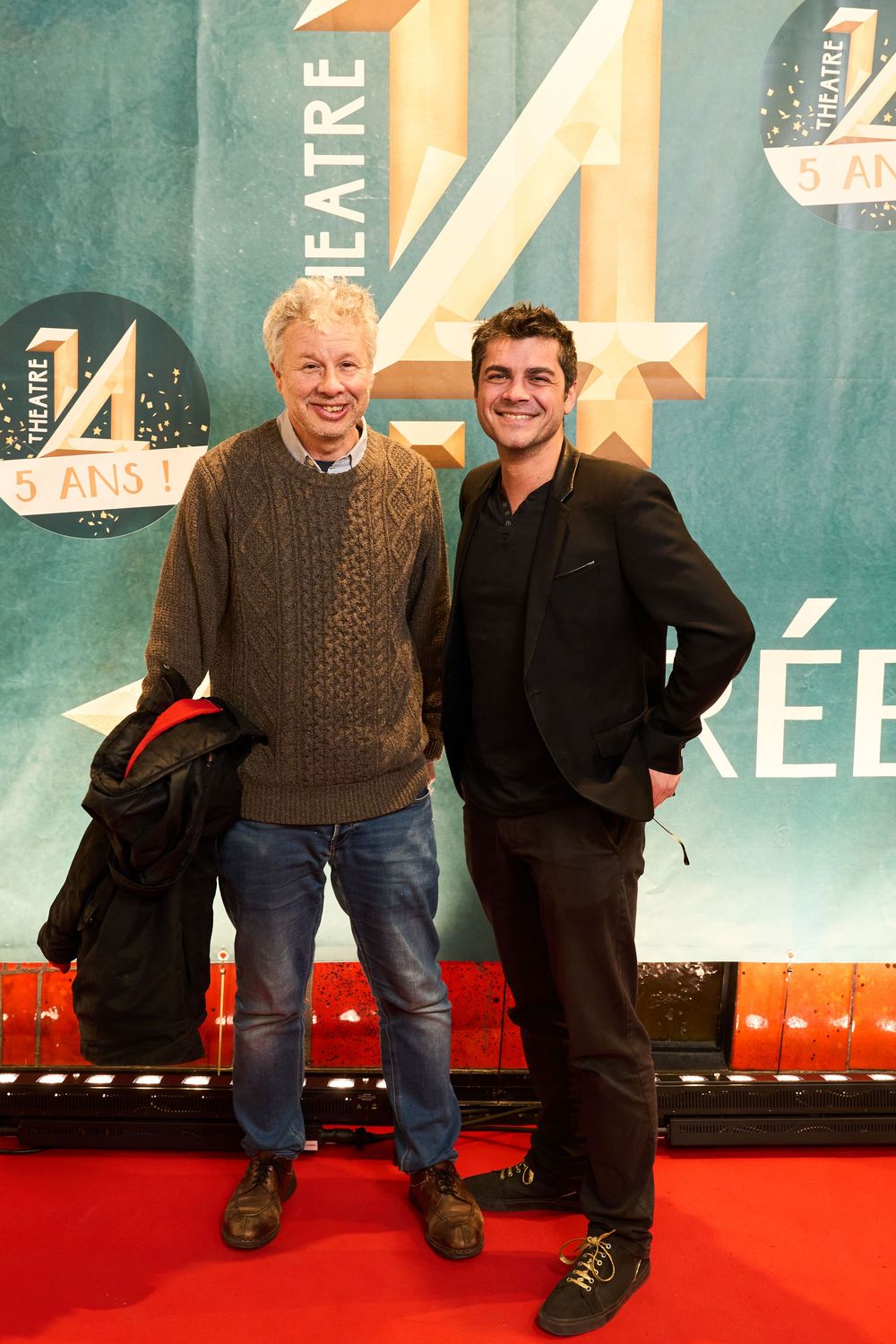 Two people posing on a red carpet in front of a teal-colored wall with large number 4 design.