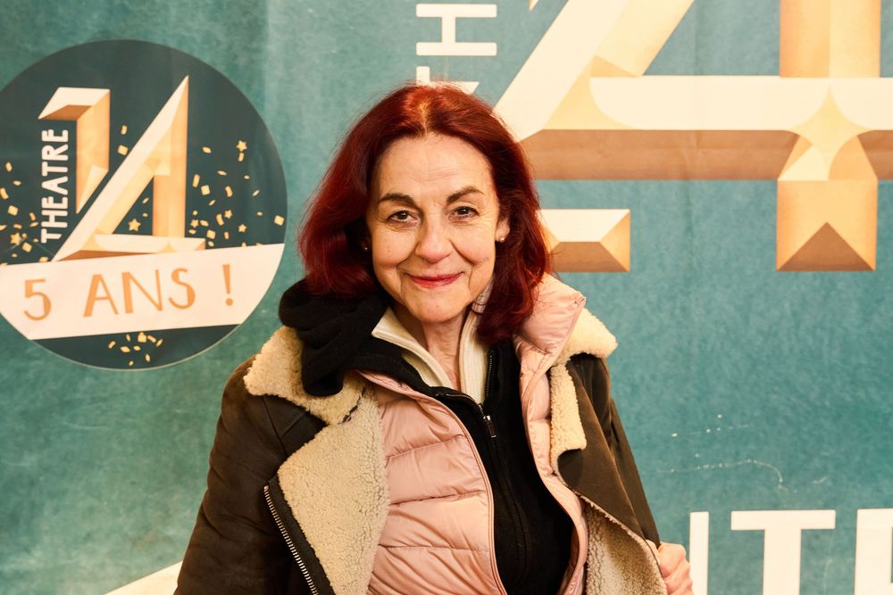 A woman wearing a beige jacket poses in front of a retro-style teal wall with decorative stars and numbers.