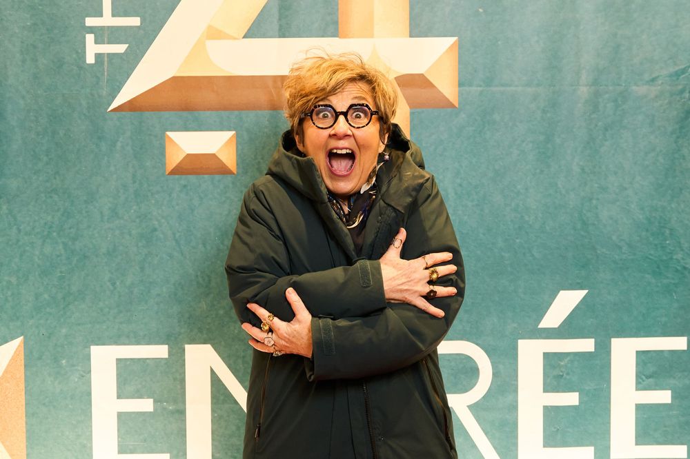 An energetic person in a black winter coat and glasses stands with arms crossed against a teal wall with geometric shapes.