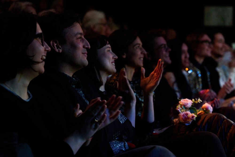 An enthusiastic audience claps and smiles while watching a performance in a darkened theater.