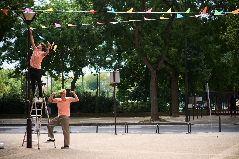 Two people work together to hang outdoor string lights in a park setting using a ladder.