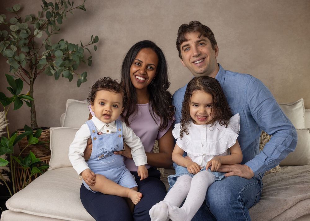 A happy family of four sits together on a light colored couch, posing warmly against a beige wall with greenery.