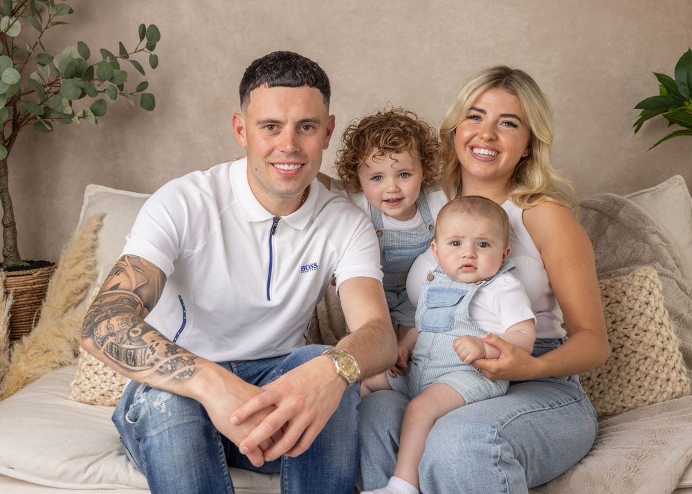 A family sits together on a beige couch in coordinated white and denim outfits for a casual indoor photoshoot.