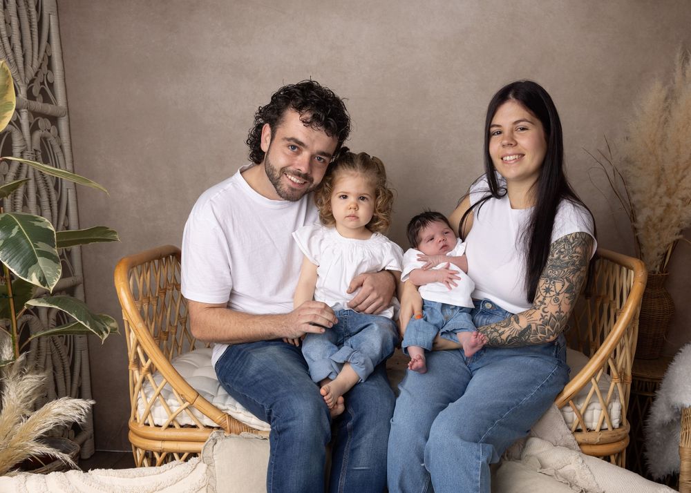A family of four sits together on a woven rattan loveseat against a neutral backdrop with dried pampas grass decor.