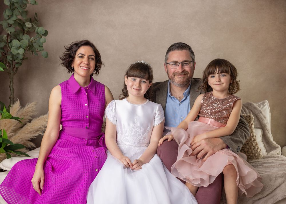 Family in coordinated outfits posing together on couch with mom in pink dress and decorative plants in background.
