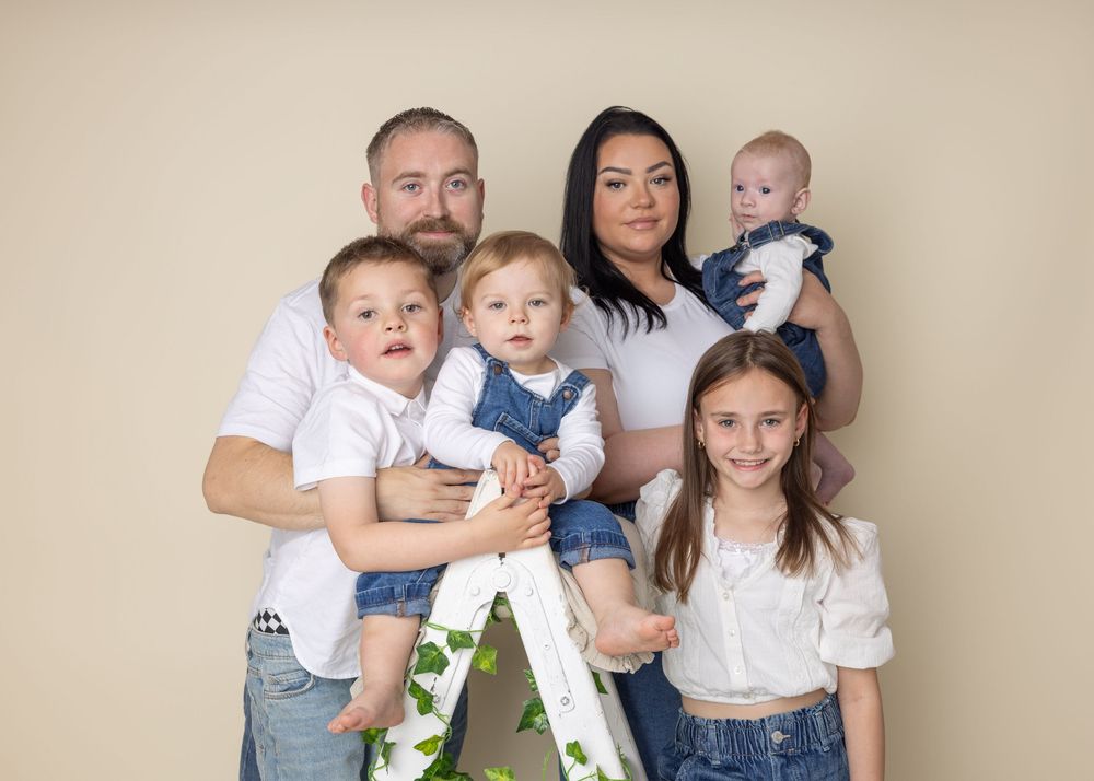 Family group in coordinated white and denim outfits poses together on a white ladder for a studio portrait.