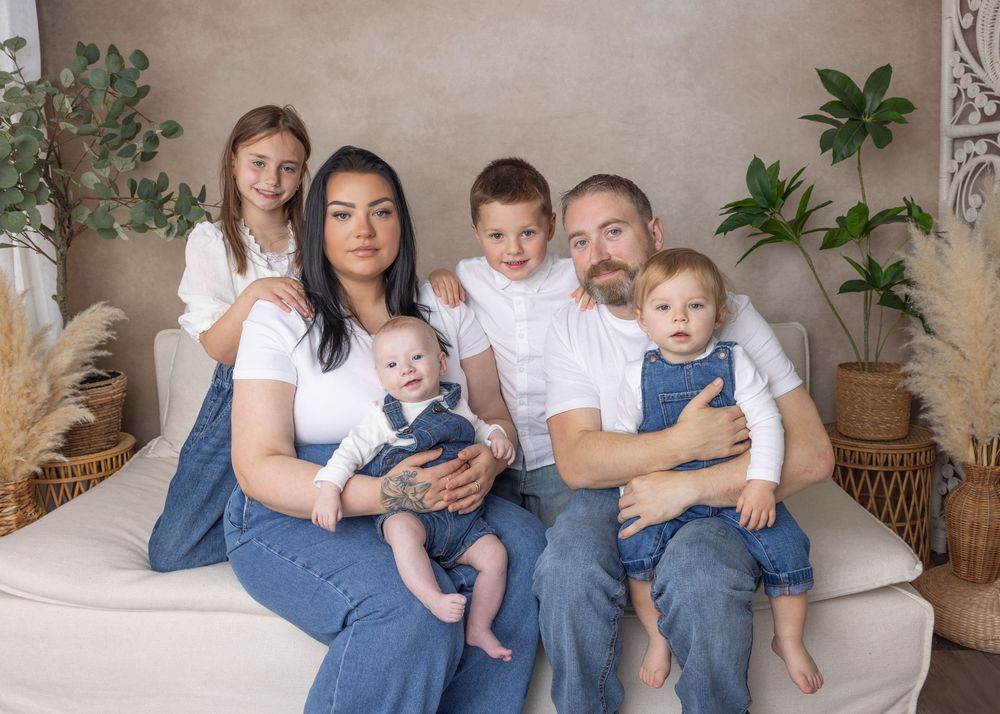 A family in coordinated denim and white outfits sits together on a light gray couch with indoor plants in the background.