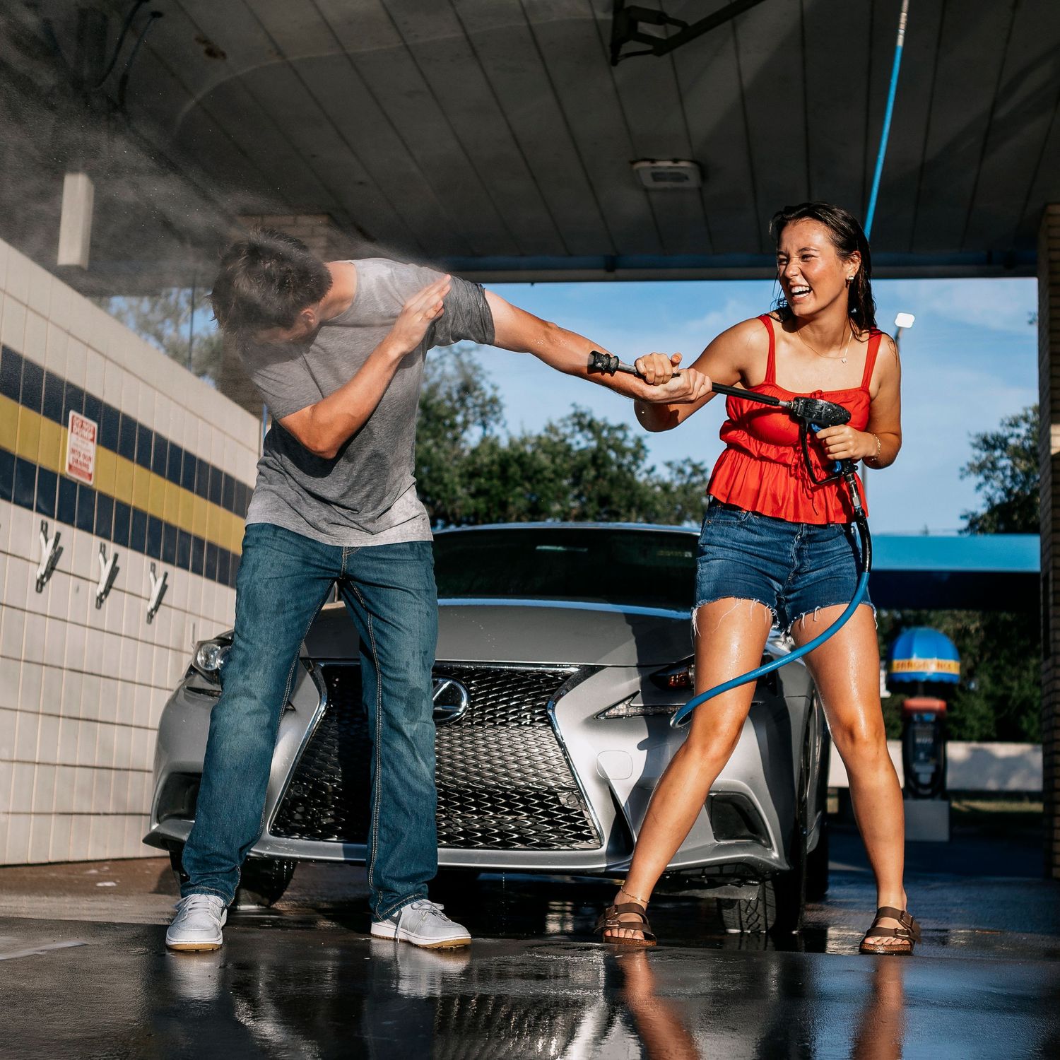 Two people having fun with water spray at a car wash location with a luxury vehicle in the background.