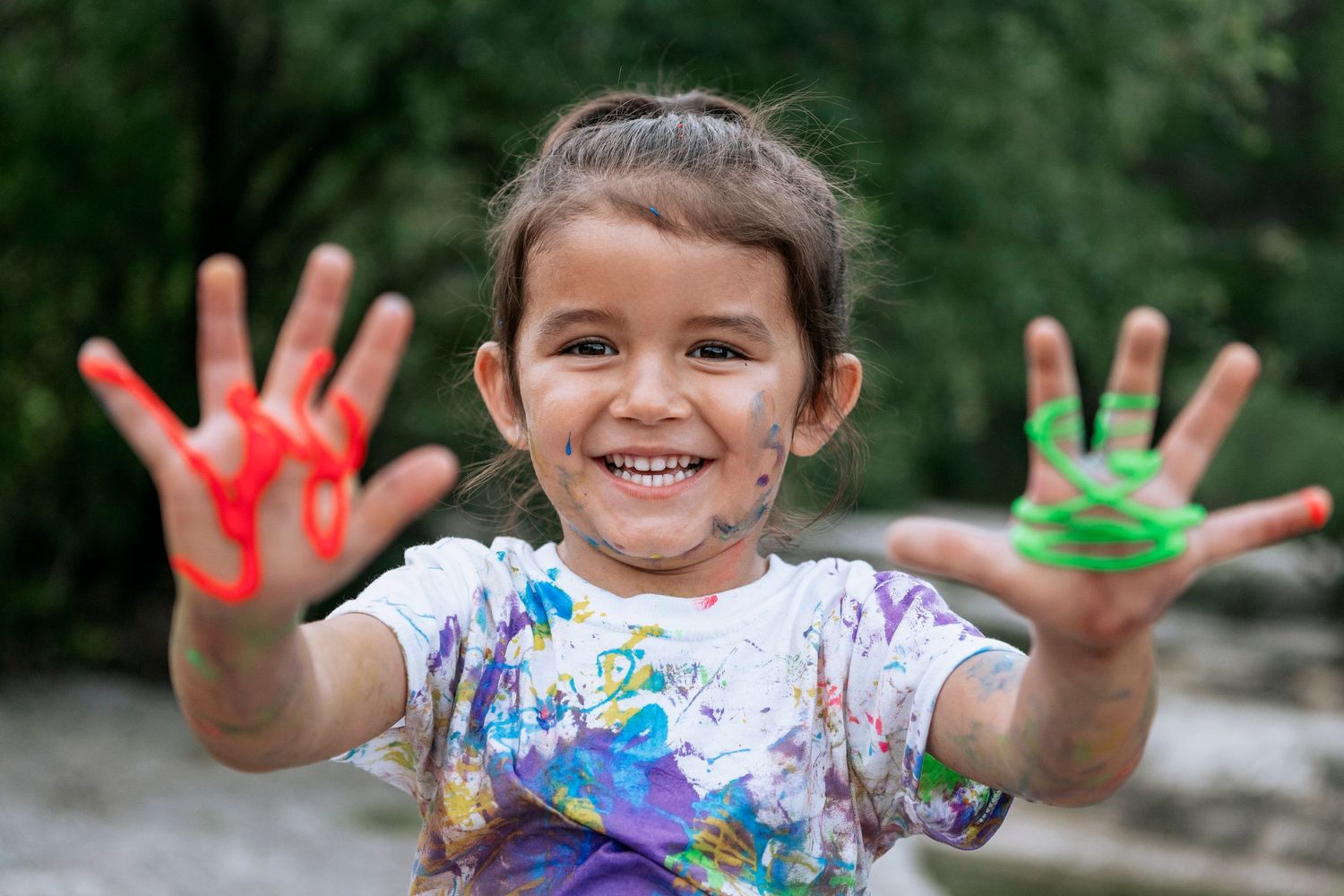 A young child shows colorful red and green paint-covered hands to camera while smiling and wearing a paint-spattered shirt.