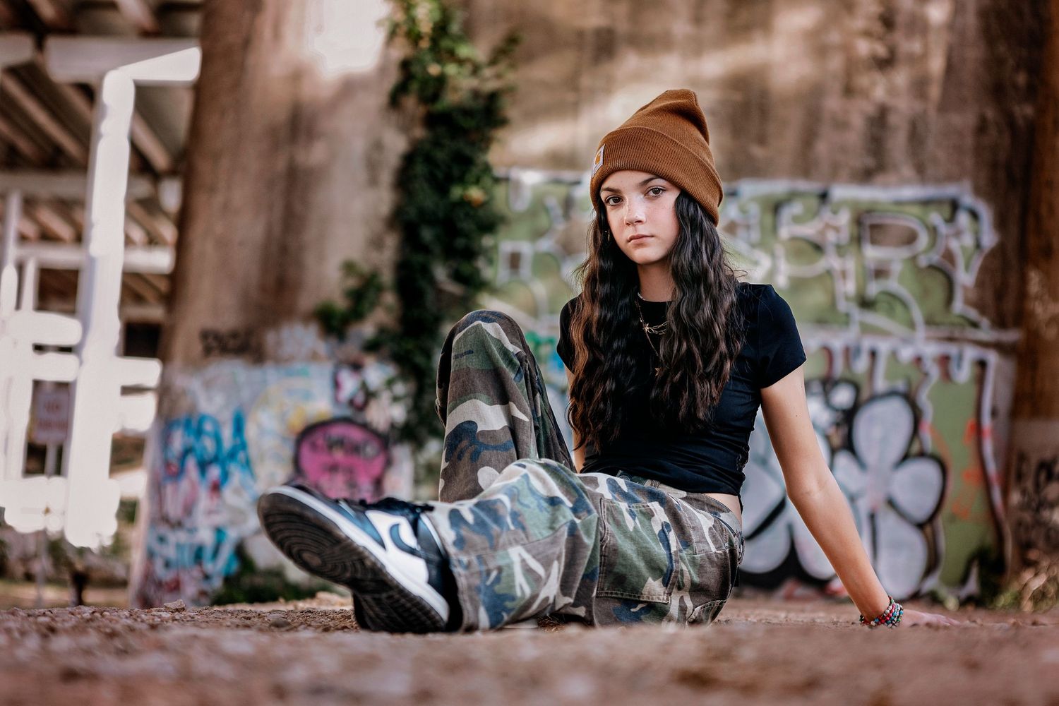 A person in casual streetwear with a brown beanie sits on urban ground with graffiti-covered walls in the background.