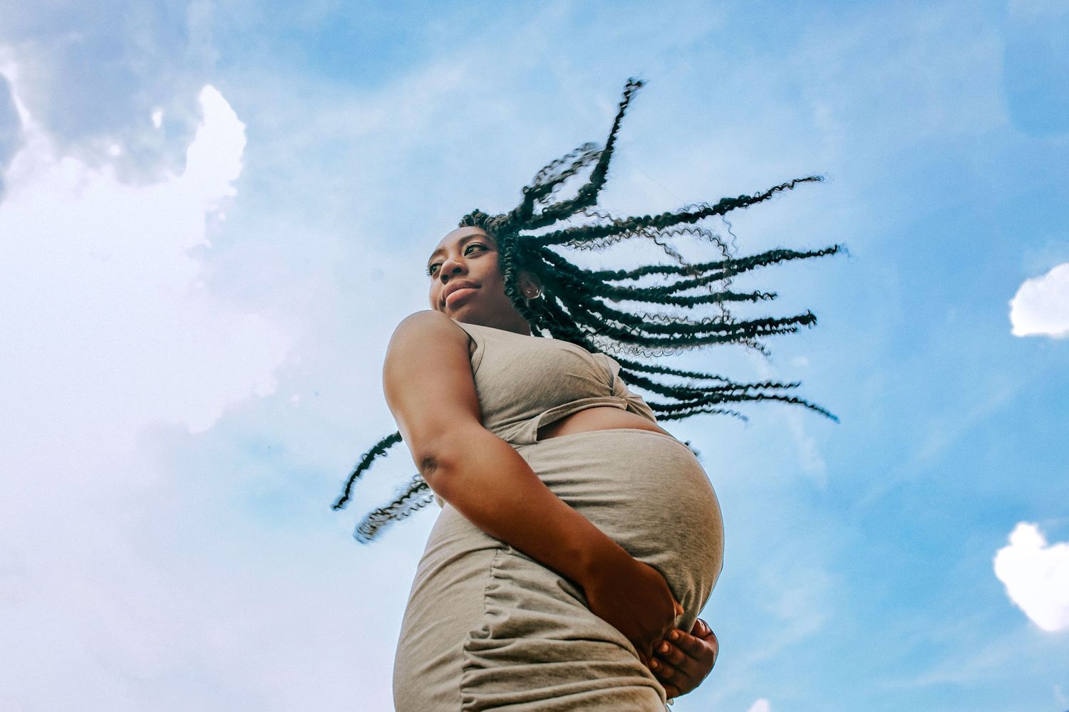 Maternity portrait of a pregnant woman with flowing braids against a blue sky, captured in an artistic, empowering photoshoot – Bedfordshire