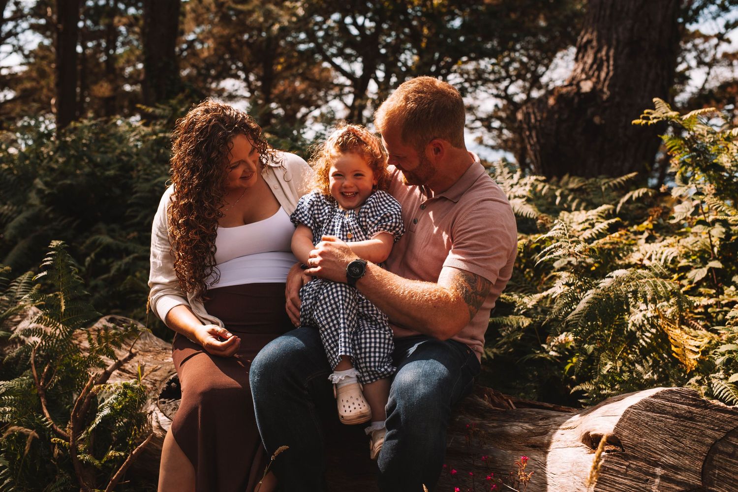 Family playing and lifting child in sunny meadow with tall grass and trees.