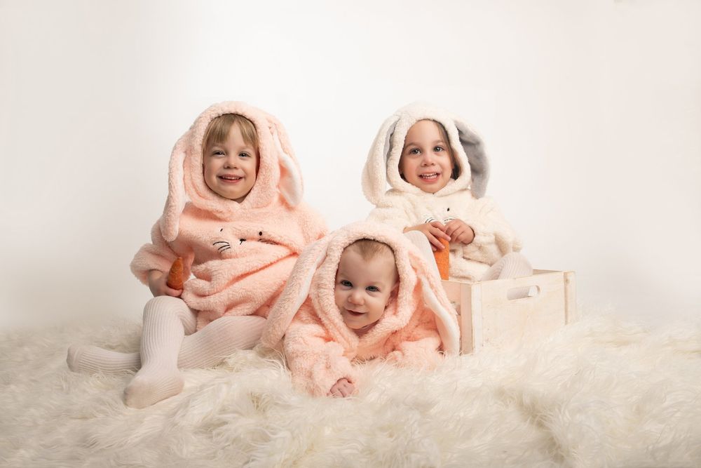 Three young children wearing soft pink hooded outfits sit together on a fluffy white rug.