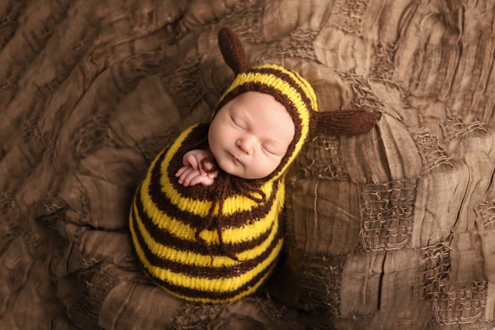 Sleeping newborn wrapped in yellow and black striped knit outfit resembling a bumblebee on brown textured backdrop.