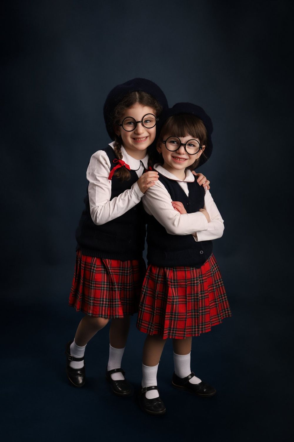 Two young students wearing matching school uniforms with plaid skirts, white shirts, and black vests pose together on dark backdrop.